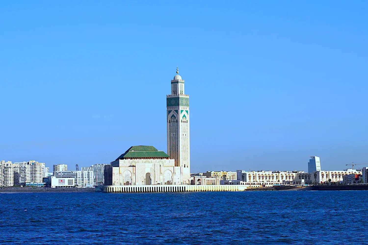 A scenic view of the Hassan II Mosque in Casablanca, Morocco, with its towering minaret, surrounded by modern city buildings and the Atlantic Ocean in the foreground