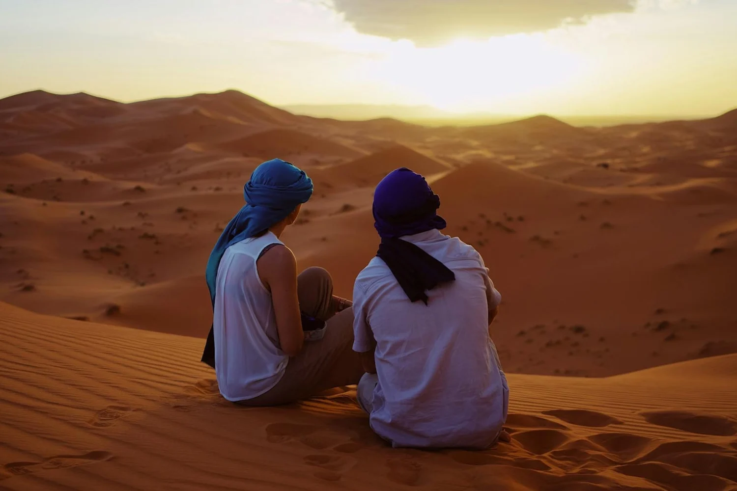 A couple sitting on golden sand dunes in Morocco, watching the sunset as the sky glows in warm hues, creating a romantic and peaceful atmosphere.