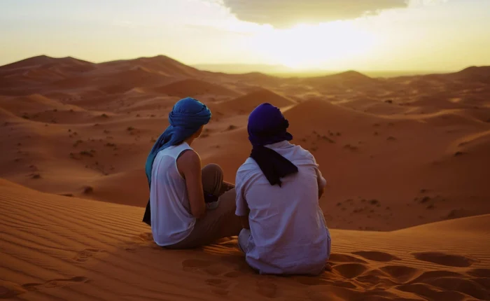 A couple sitting on golden sand dunes in Morocco, watching the sunset as the sky glows in warm hues, creating a romantic and peaceful atmosphere.