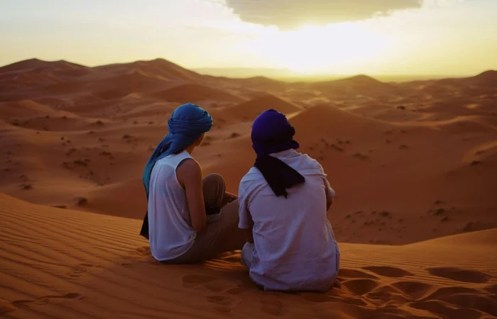 A couple sitting on golden sand dunes in Morocco, watching the sunset as the sky glows in warm hues, creating a romantic and peaceful atmosphere.
