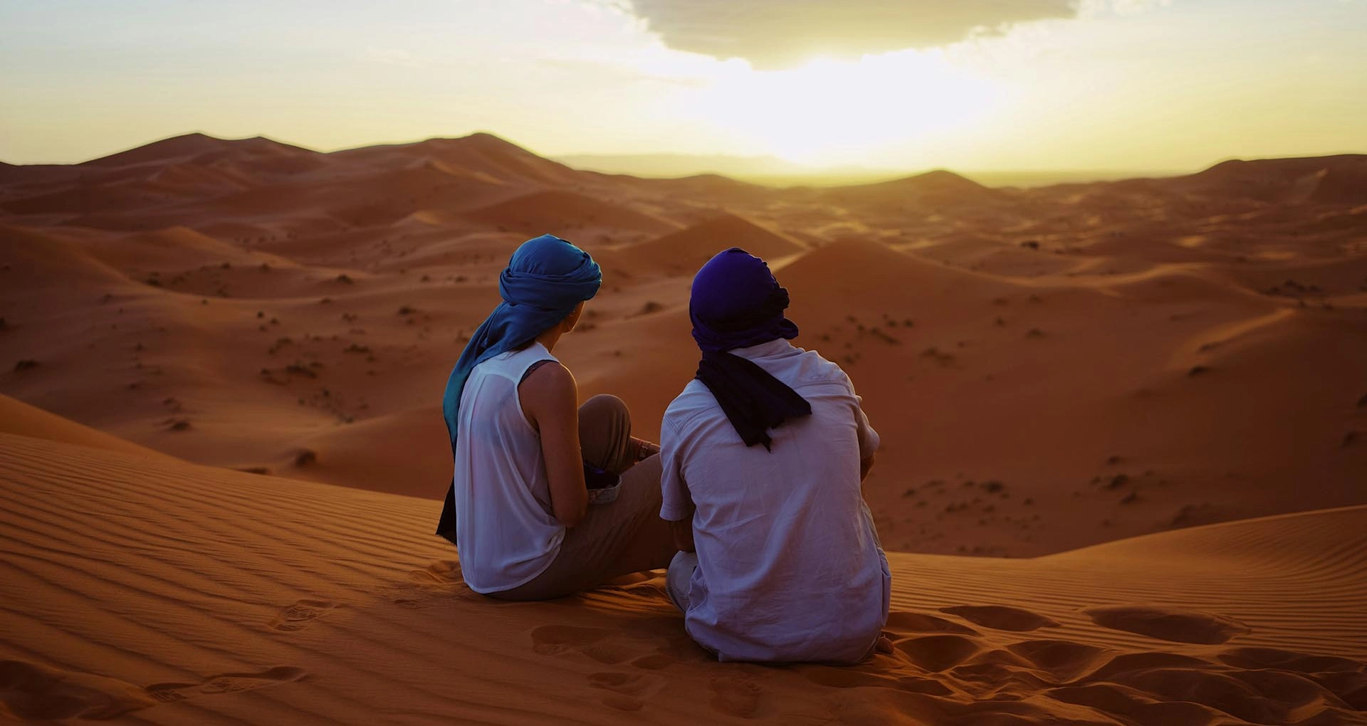 A couple sitting on golden sand dunes in Morocco, watching the sunset as the sky glows in warm hues, creating a romantic and peaceful atmosphere.